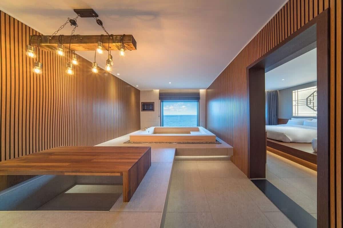 A modern spa area with a wooden wall, a wooden bench, a small seating area, and a view of a bedroom with a bed and a window.