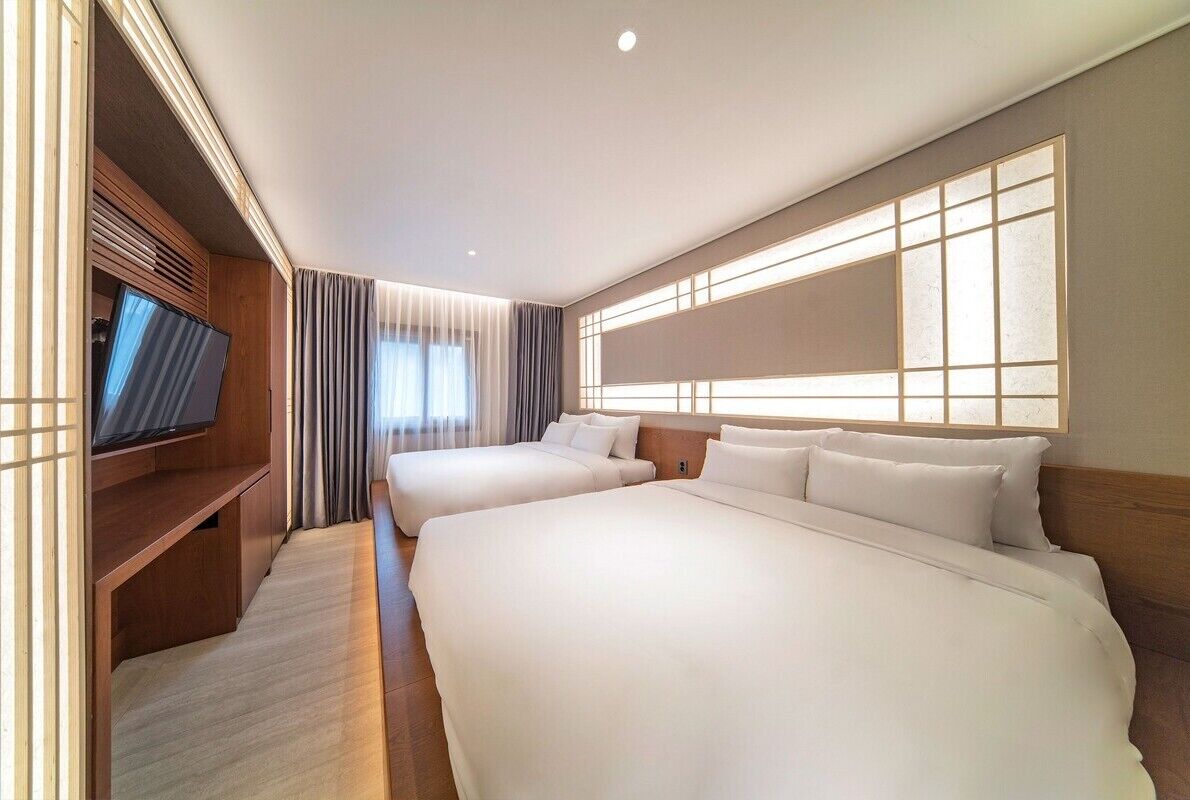 A hotel room with two beds, a TV, and a large window with blinds.