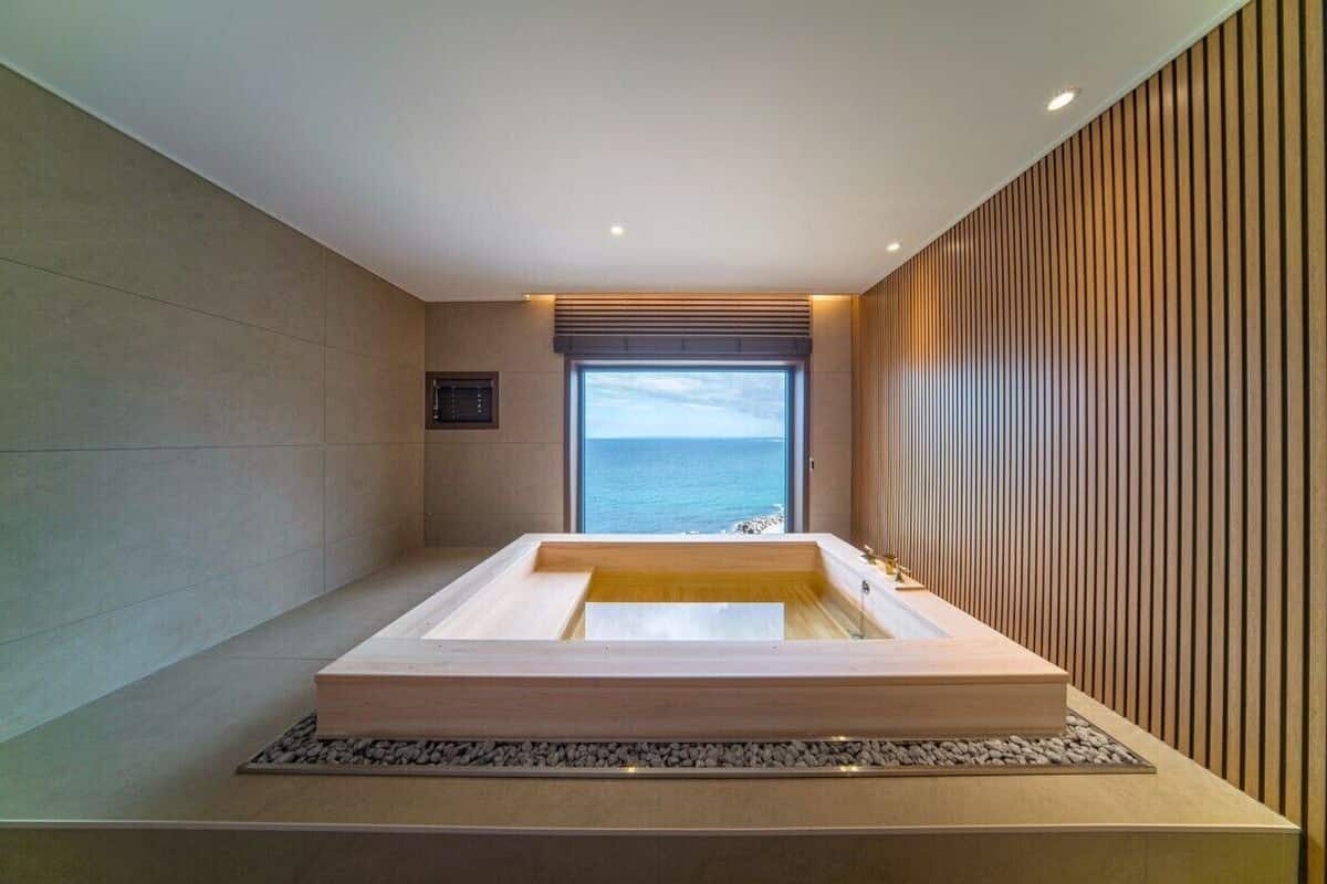 A modern bathroom with a large bathtub, a view of the sea, and a wooden panel wall.