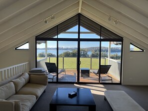 A spacious room with a large sliding glass door leading to an outdoor deck. Inside, there is a beige sofa, a black coffee table, and two chairs. - Black Bream Point Cabins (North Narooma)