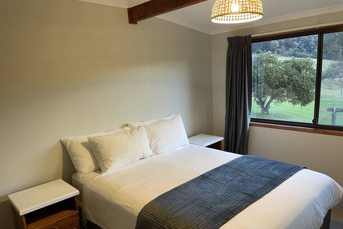 A bedroom with a bed, a nightstand, a wooden drawer unit, a window with a view of greenery, and a hanging light fixture.