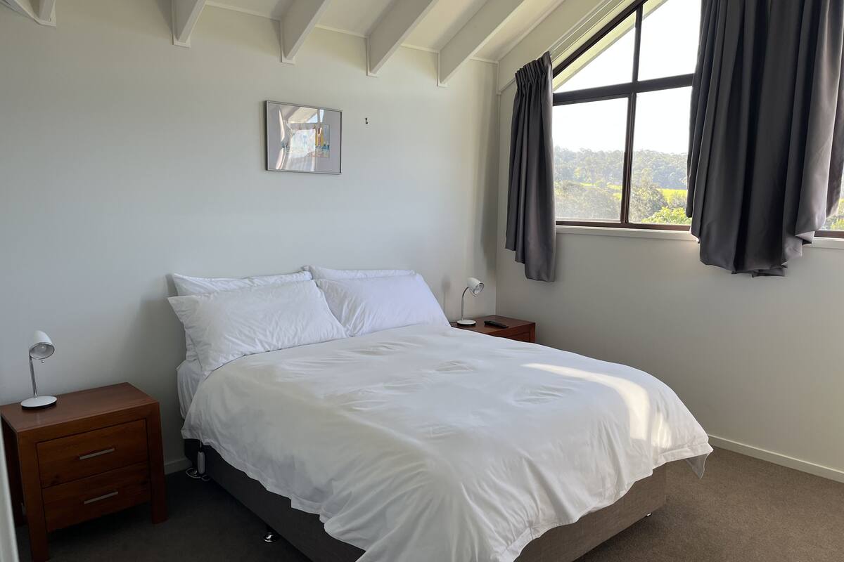 A bedroom with a large bed, two bedside tables, a nightstand, a window with curtains, and a framed picture on the wall.