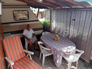 Een overdekt terras met een tafel, stoelen, een camper en een decoratieve bloemstelling.