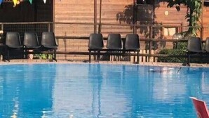 Área de piscina com cadeiras de plástico vermelhas e uma mesa, rodeada por um pátio de tijolos.