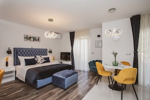 Quarto moderno com cama grande, mesas de cabeceira, escrivaninha, espaço de refeições com cadeiras e televisão.
