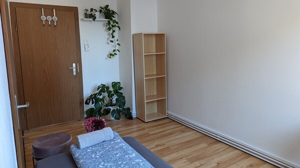 Quarto com porta de madeira, cama com colcha estampada, um pequeno banquinho, uma planta alta e uma estante de madeira.