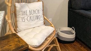 Een rieten stoel met een kussen waarop staat: 'THE BEACH IS CALLING'.