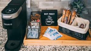 Een keuken met witte kastjes, een koffiezetapparaat, een dienblad met koffiekapsules, een bordje met de tekst 'BUT FIRST' en een mand met snacks.