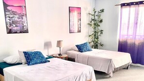 Habitación de hotel con dos camas individuales, una mesita, una lámpara y un ventanal grande con cortinas.