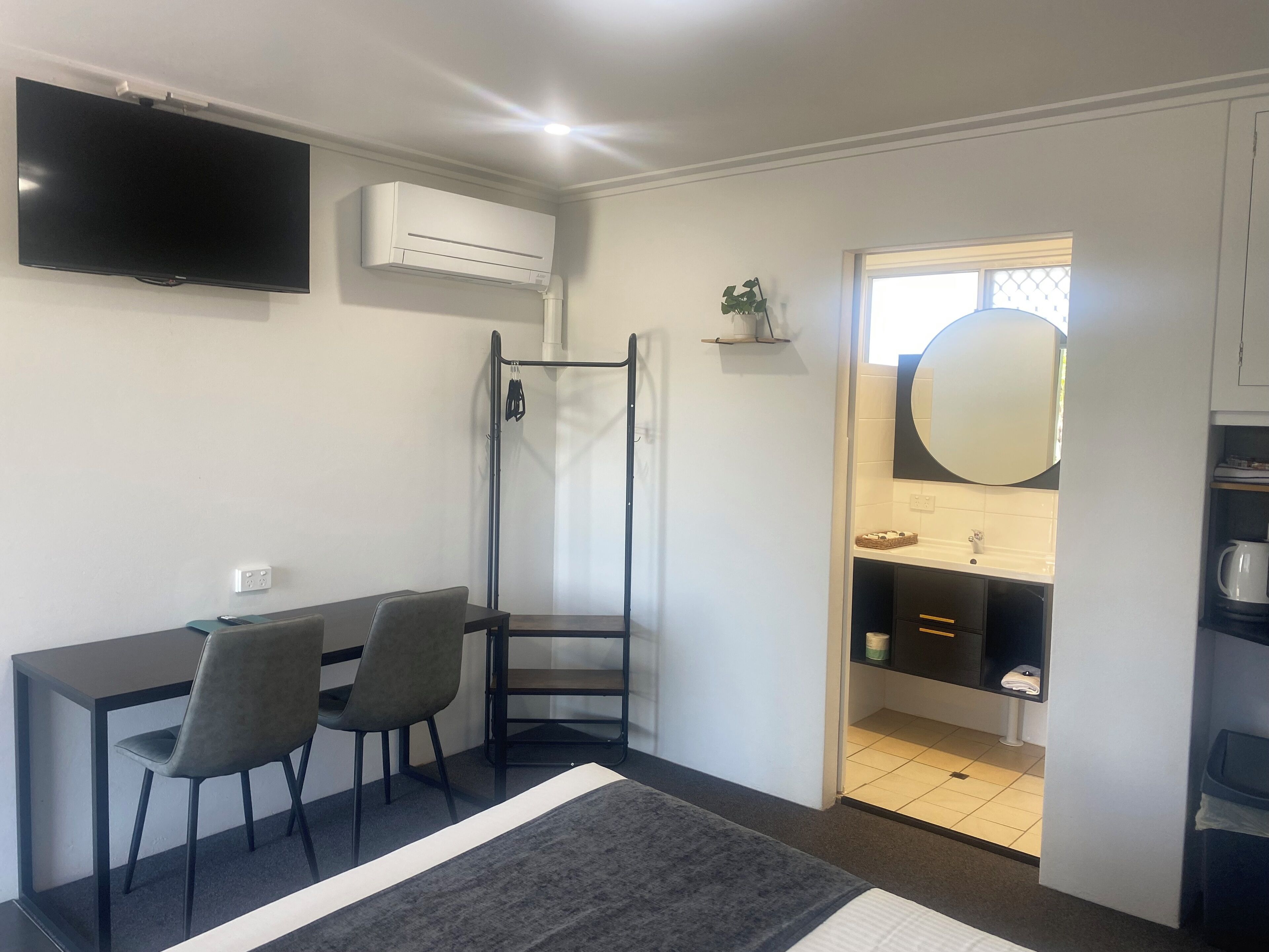 A compact room with a bed, a desk with two chairs, a TV, an air conditioner, a small shelf with a plant, a mirror, and a bathroom visible through an open door.