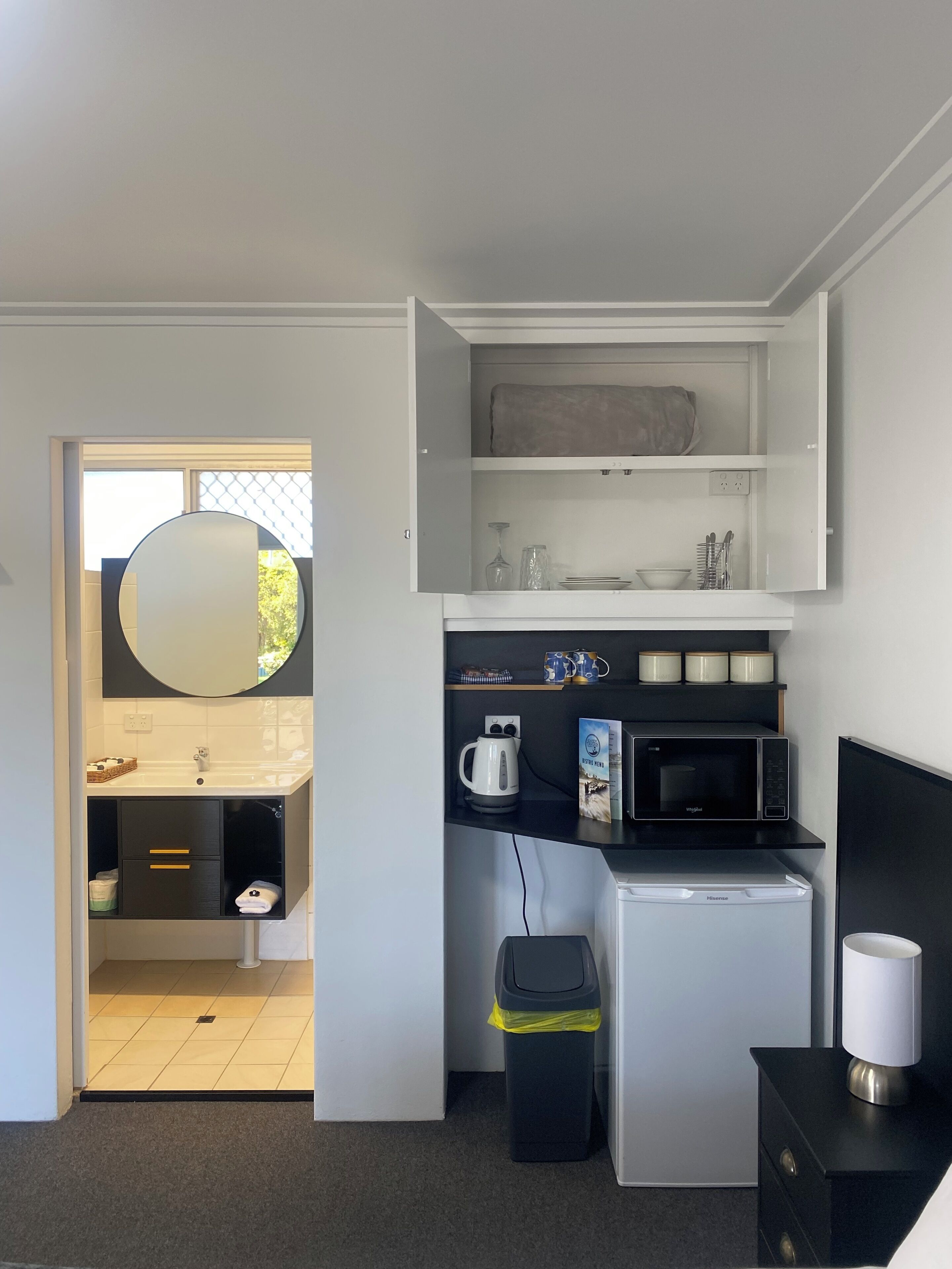 A compact kitchenette with a microwave, kettle, and a small refrigerator. There is a trash bin and a cabinet with drawers. A mirror is mounted above the sink.