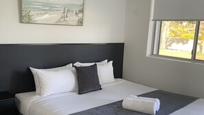 Un lit bien fait, avec du linge de lit blanc et gris, une tête de lit sombre et un tableau représentant une scène de plage accroché au mur.