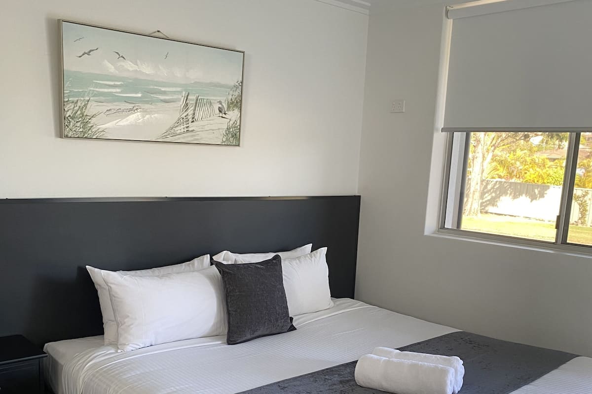 A neatly made bed with white and gray linens, a dark headboard, and a painting of a beach scene on the wall.
