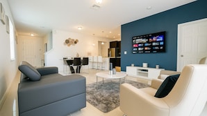 A modern living room with a grey sofa, a white sofa, and a coffee table. There is a TV mounted on the wall and a dining area in the background. - Stylish Kissimmee Getaway Game Room – 99 Dollar Deal! (Kissimmee)
