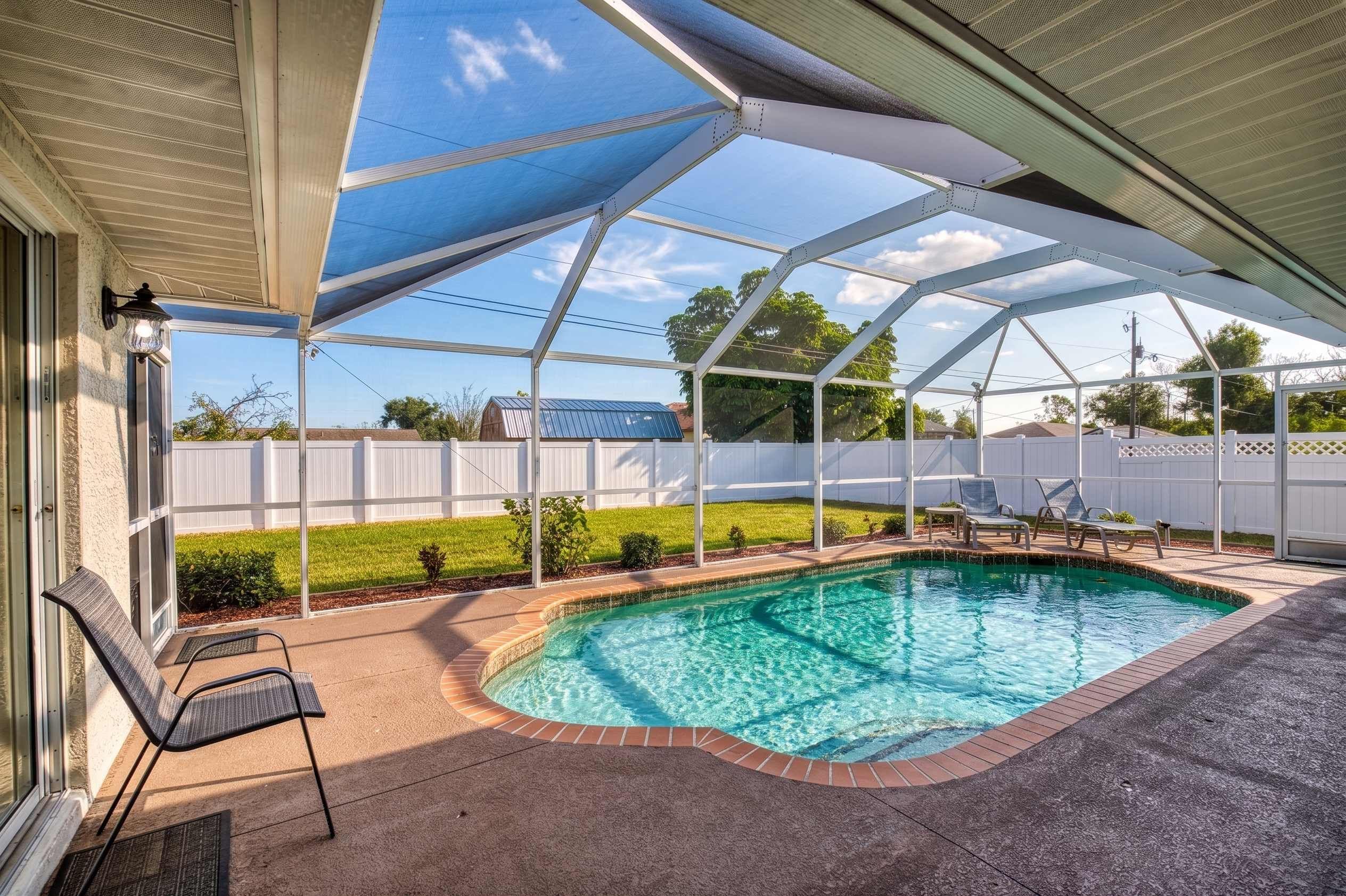 Una piscina exterior techada con un elemento acuático de agua azul cristalina, rodeada por una zona de terraza con asientos y una cerca blanca al fondo.