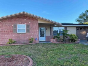 A single-story brick house with a front porch, a garage, and a small front yard with plants. - Sunrise, Suntan, Sunset repeat! Coastal getaway! 2 bd, pet friendly, fenced yard (Holiday)