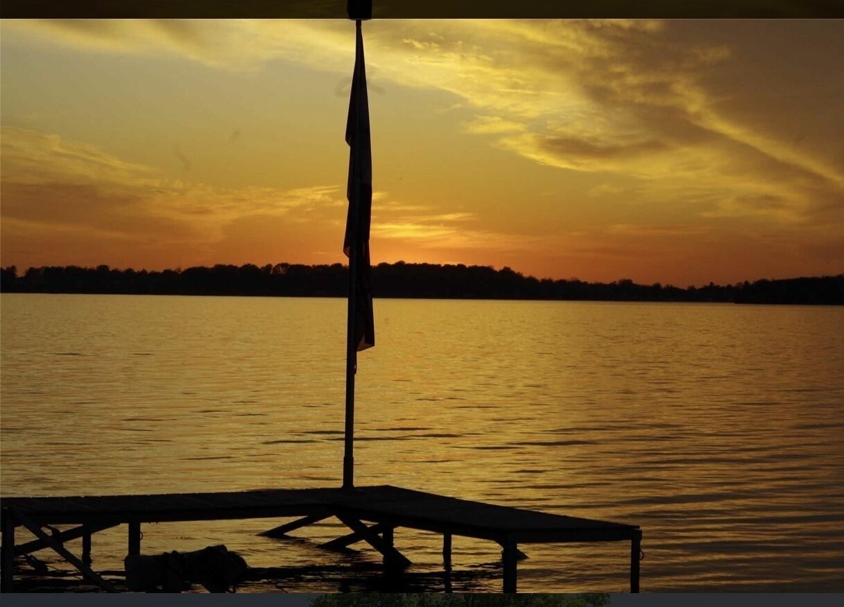 Un coucher de soleil sur un lac paisible, avec un quai et un mât de drapeau.