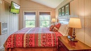 A bedroom with a large bed, two bedside lamps, a television mounted on the wall, and a window with a view of greenery.