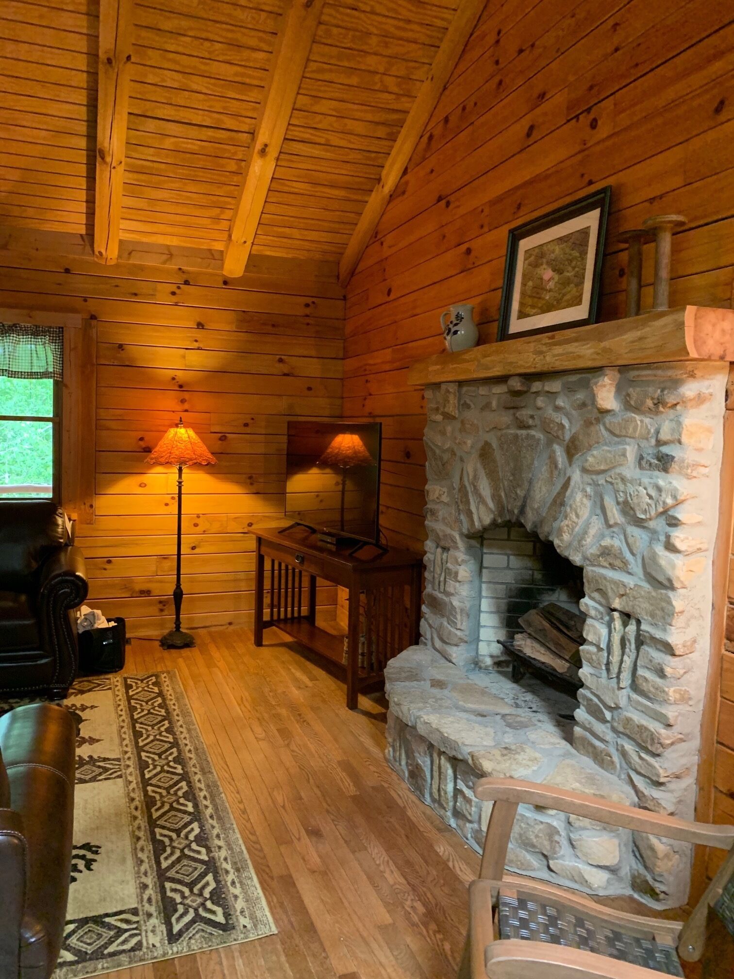 A cozy log cabin interior with a stone fireplace, wooden walls, a wooden ceiling, a patterned rug, and a leather chair.