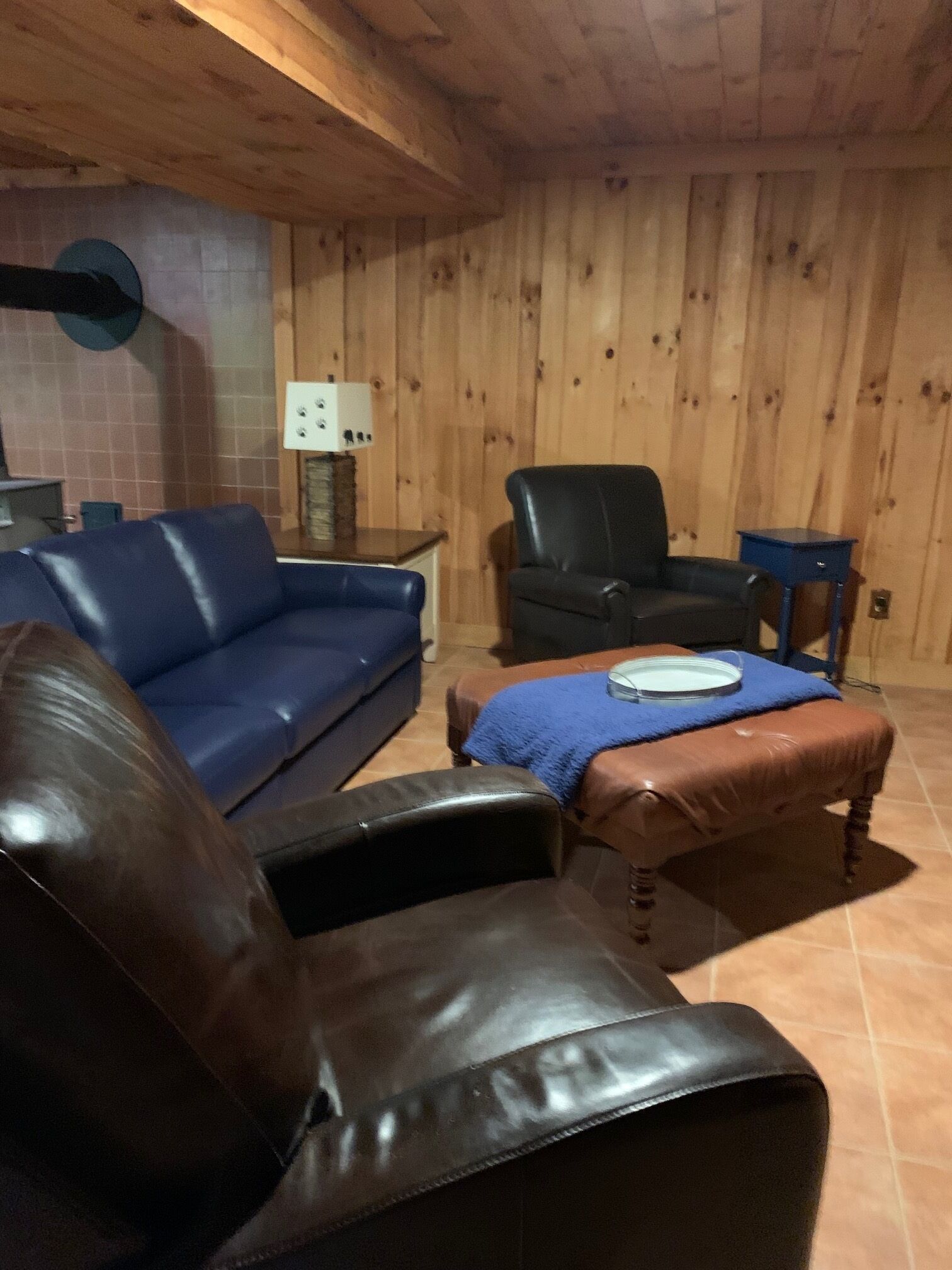 A room with a wooden paneled wall, a blue sofa, a black leather armchair, and a small table with a blue cloth.