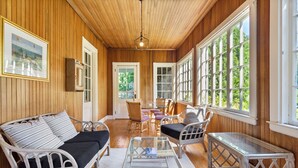 Una habitación acogedora con techo de madera, un sofá de mimbre blanco con cojines negros, una mesa de centro de vidrio y una mesita auxiliar de mimbre.