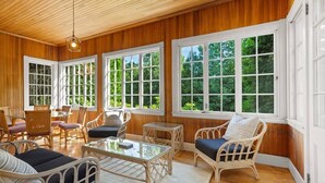 Una habitación luminosa con paredes de madera, ventanas amplias, mobiliario de mimbre y una mesa de centro de vidrio.