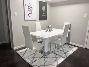 Een eethoek met een witte tafel en stoelen, een kamerbreed tapijt met dessin en twee Gucci posters aan de muur.