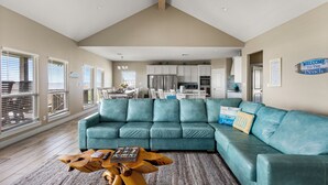 A spacious living room with a teal sectional sofa, a wooden coffee table, and a kitchen in the background. - Beachfront | Firepit, Volleyball & Gazebo Dining (Port Bolivar)