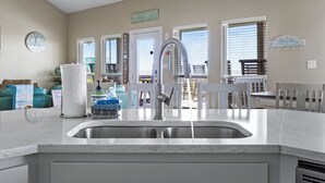 A kitchen with a sink, a faucet, and a countertop. There are chairs and a table in the background. - Beachfront | Firepit, Volleyball & Gazebo Dining (Port Bolivar)
