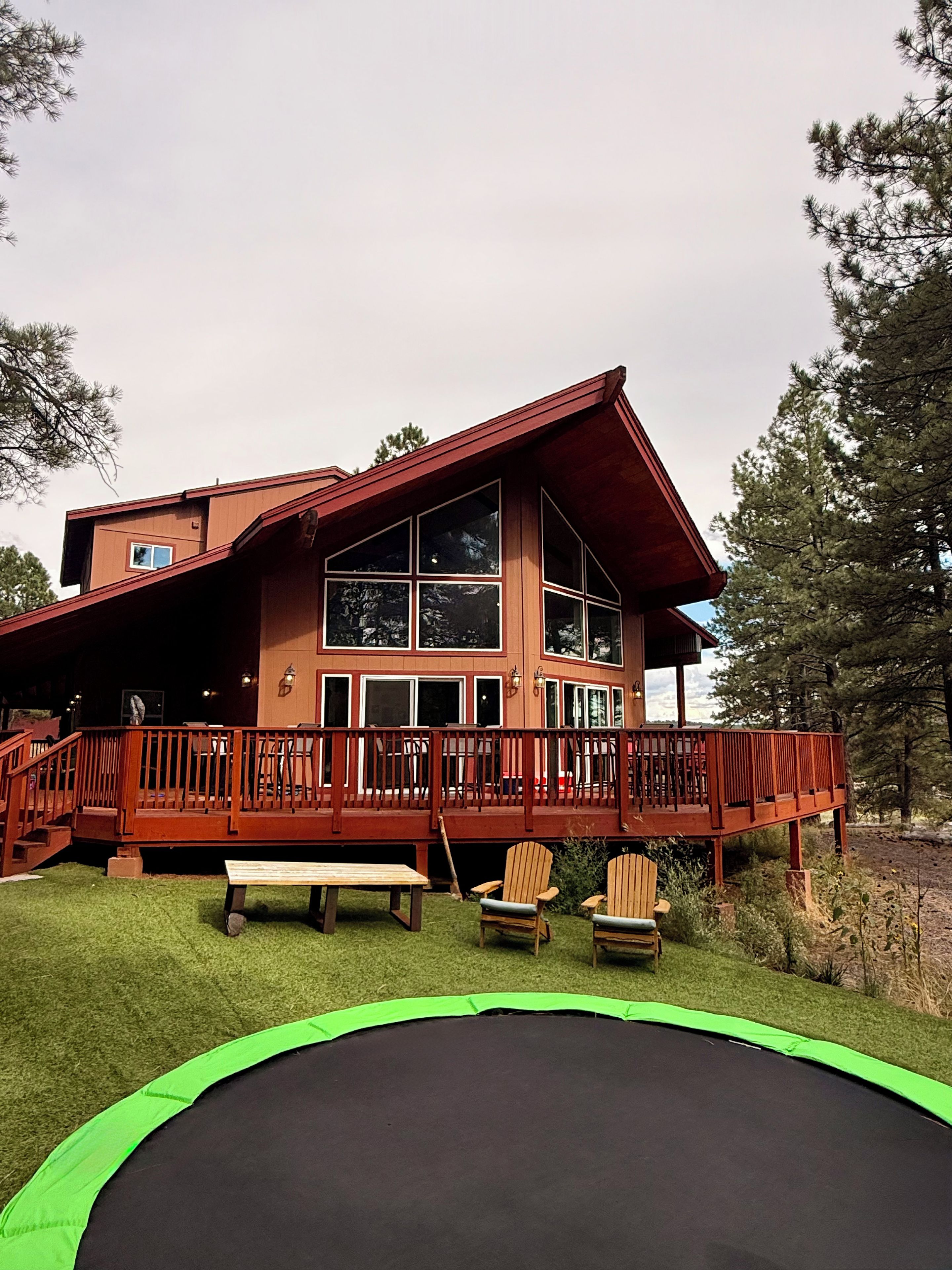 A two-story wooden house with a balcony, a trampoline, and two chairs.