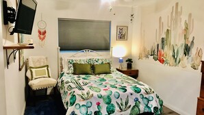 A bedroom with a bed, a chair, a desk, a wall-mounted TV, and a cactus-themed wall art. - Roadrunner Bedroom in La Quinta Cove (La Quinta)