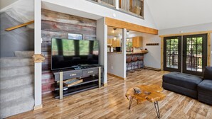 Een ruime woonkamer met een televisie, houten vloer, een bank en een trap naar een verdieping erboven.