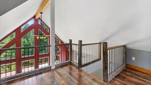 Ruime zolderkamer met schuine balkenplafond, houten balken en een raam met rode omlijsting dat uitkijkt op een balkon.