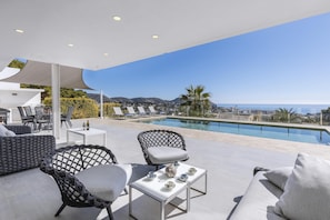 Uma área de estar moderna ao ar livre, com piscina, apresentando uma mesa branca, duas cadeiras e um sofá.