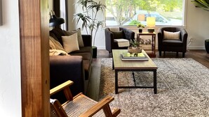 Un salon comprenant un canapé, deux fauteuils, une table basse et une chaise en bois. Une grande fenêtre offre une vue sur les arbres et une voiture garée à l’extérieur.