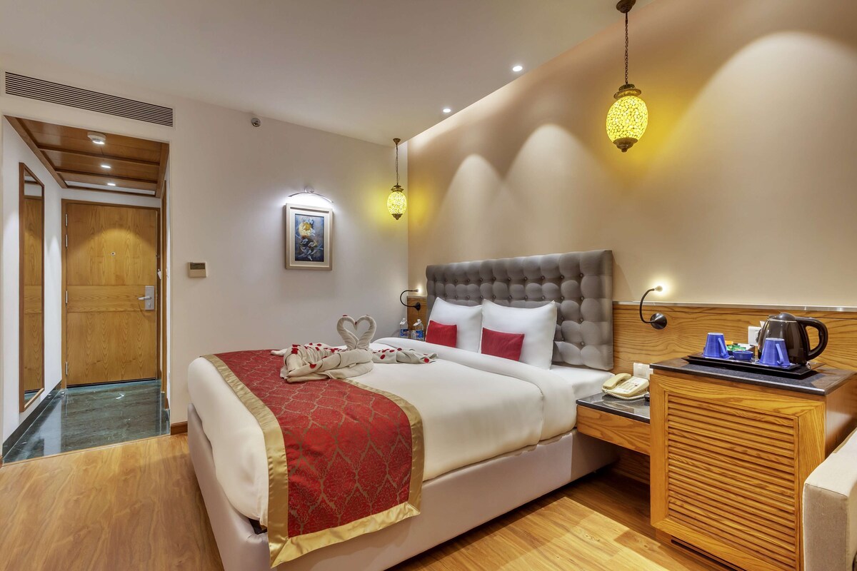 A hotel room with a large bed, a wooden nightstand, a small table with a kettle, and a wall-mounted telephone.