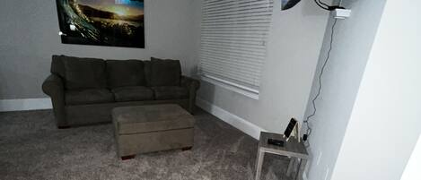 A living room with a sectional sofa, a small ottoman, a flat-screen TV mounted on the wall, and a window with blinds.