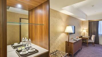 A hotel room with a wooden wardrobe, a glass shelf, a desk with a chair, a TV, and a lamp.