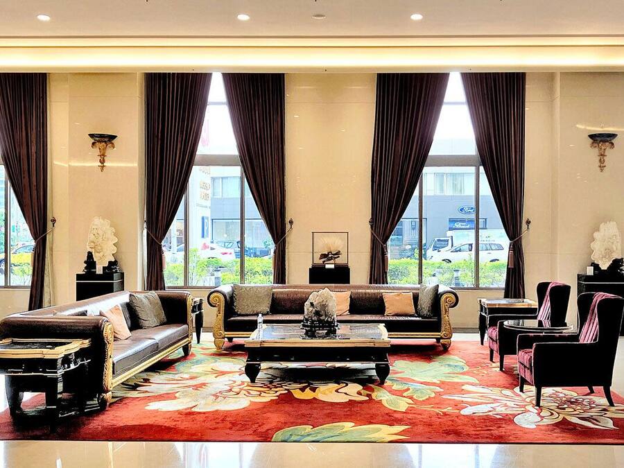 A spacious living room with a sofa set, a coffee table, and a red floral rug. Large windows with curtains allow natural light and views of the outside.