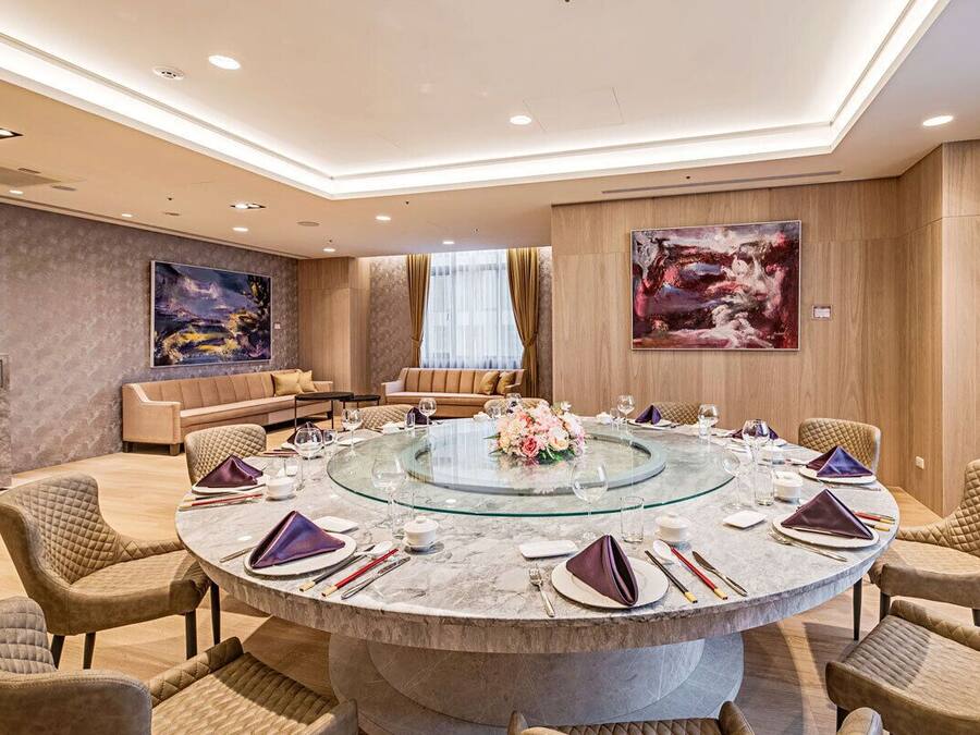 Una mesa de comedor redonda con tapa de vidrio, rodeada de sillas acolchadas, en una habitación luminosa con un sofá y obras de arte en las paredes.