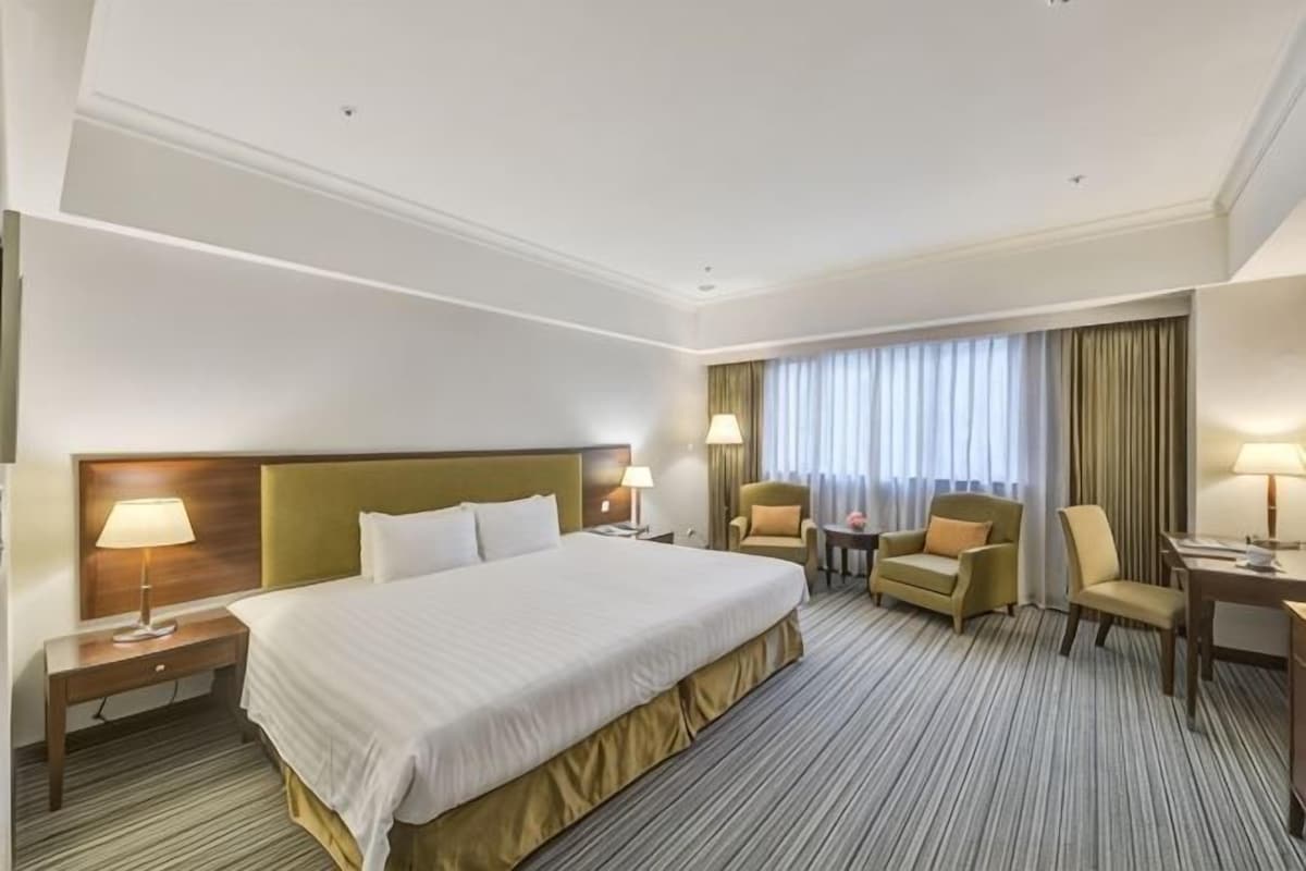 Quarto de hotel com uma cama grande, duas poltronas, uma mesa pequena e uma escrivaninha com abajur.