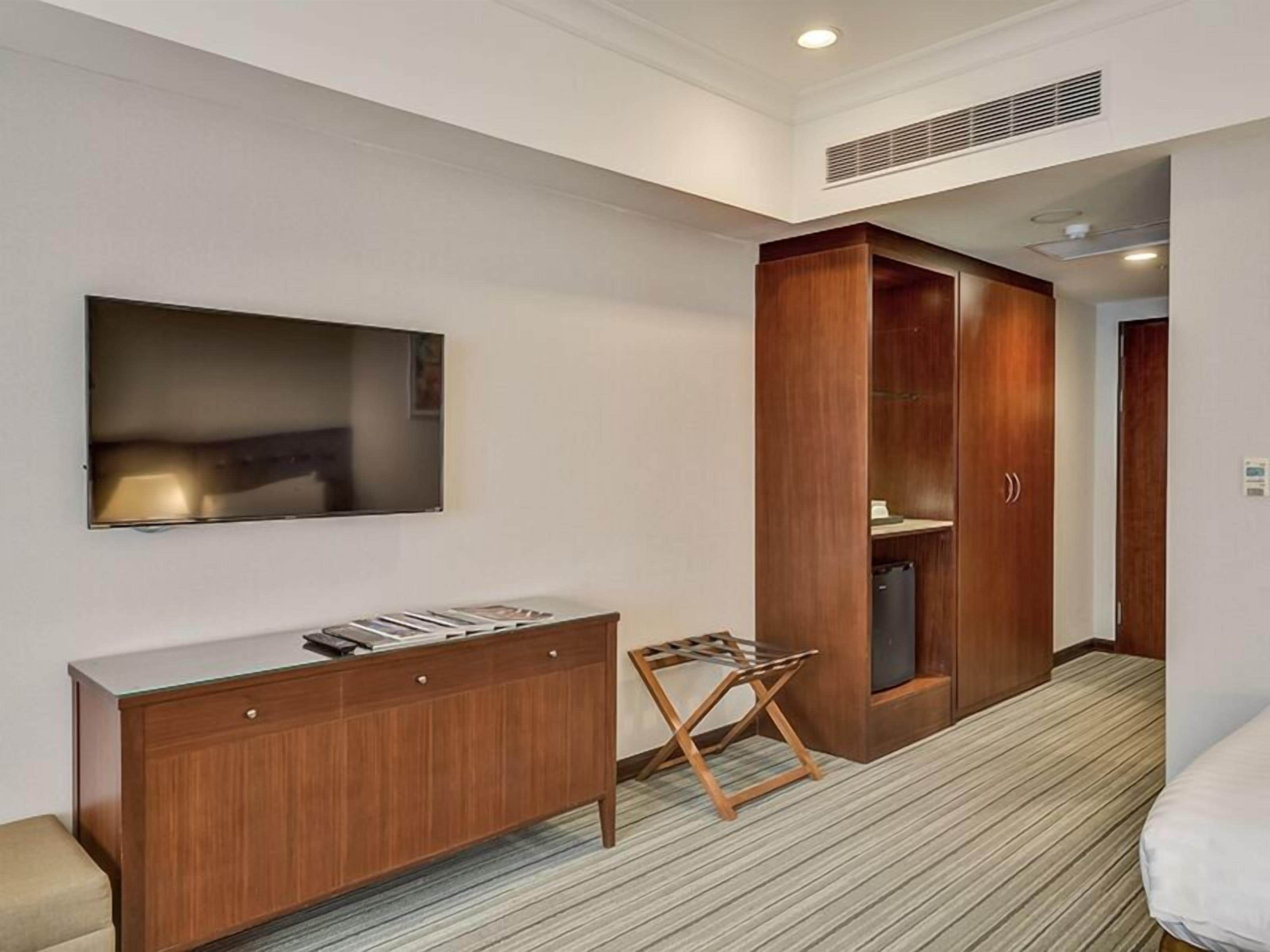 A hotel room with a wooden wardrobe, a flat-screen TV mounted on the wall, a small wooden stool, and a bed.
