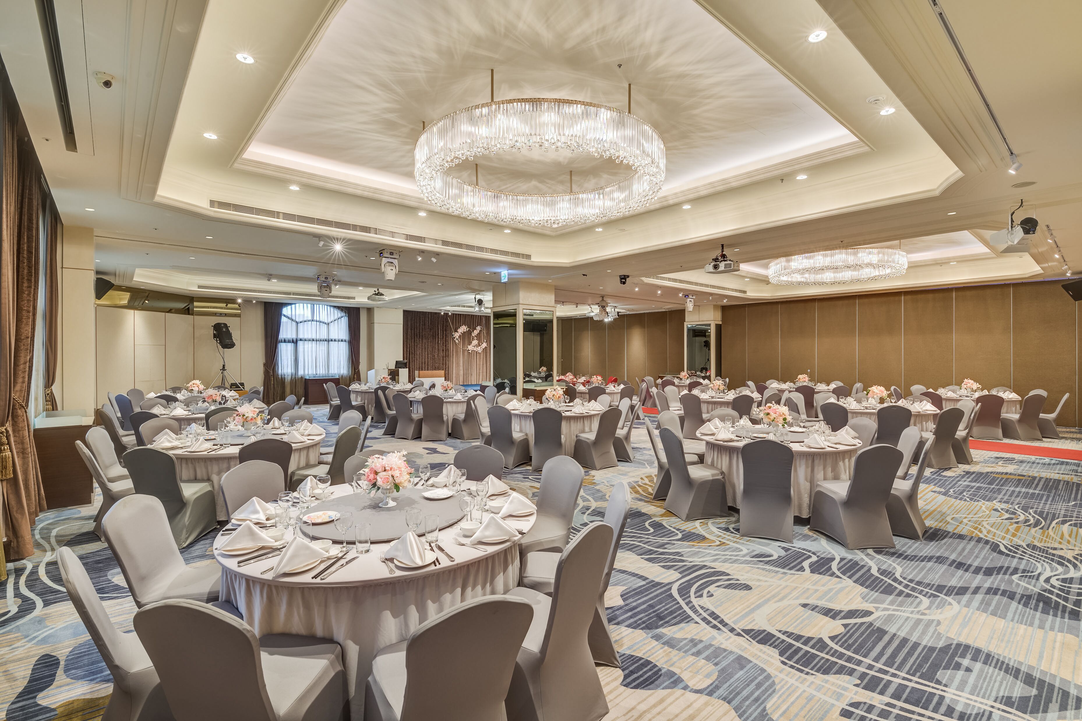 Un salón de banquetes con mesas redondas dispuestas para cenar, grandes candelabros y una alfombra estampada.