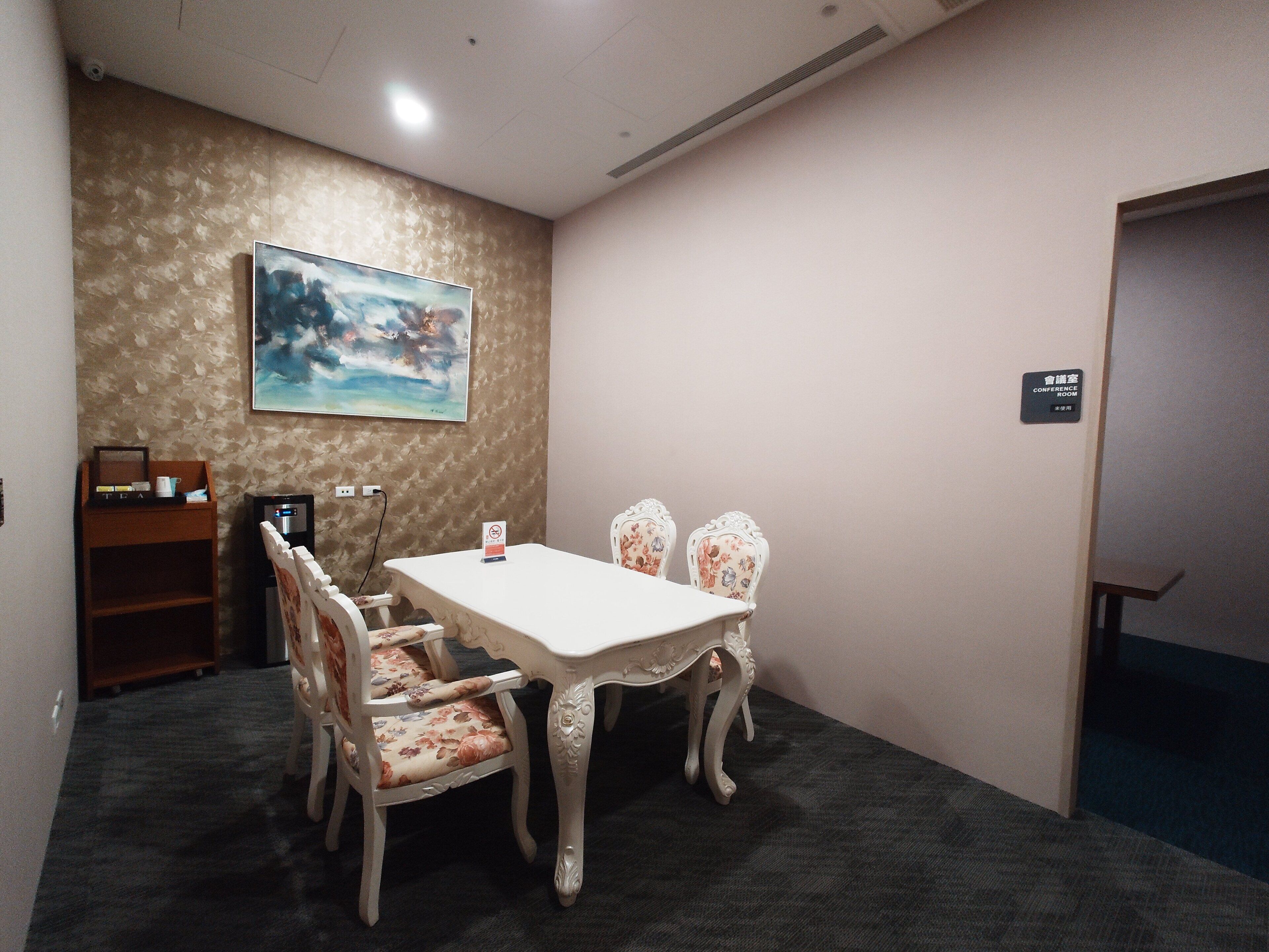 Un salón de conferencias con una mesa blanca y sillas con estampado floral, un cuadro en la pared y un cartel que indica que es una sala de conferencias.