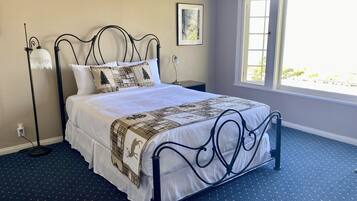 A neatly made bed with a white comforter and decorative pillows, a black metal headboard, a bedside table with a lamp, and a window with a view of greenery.