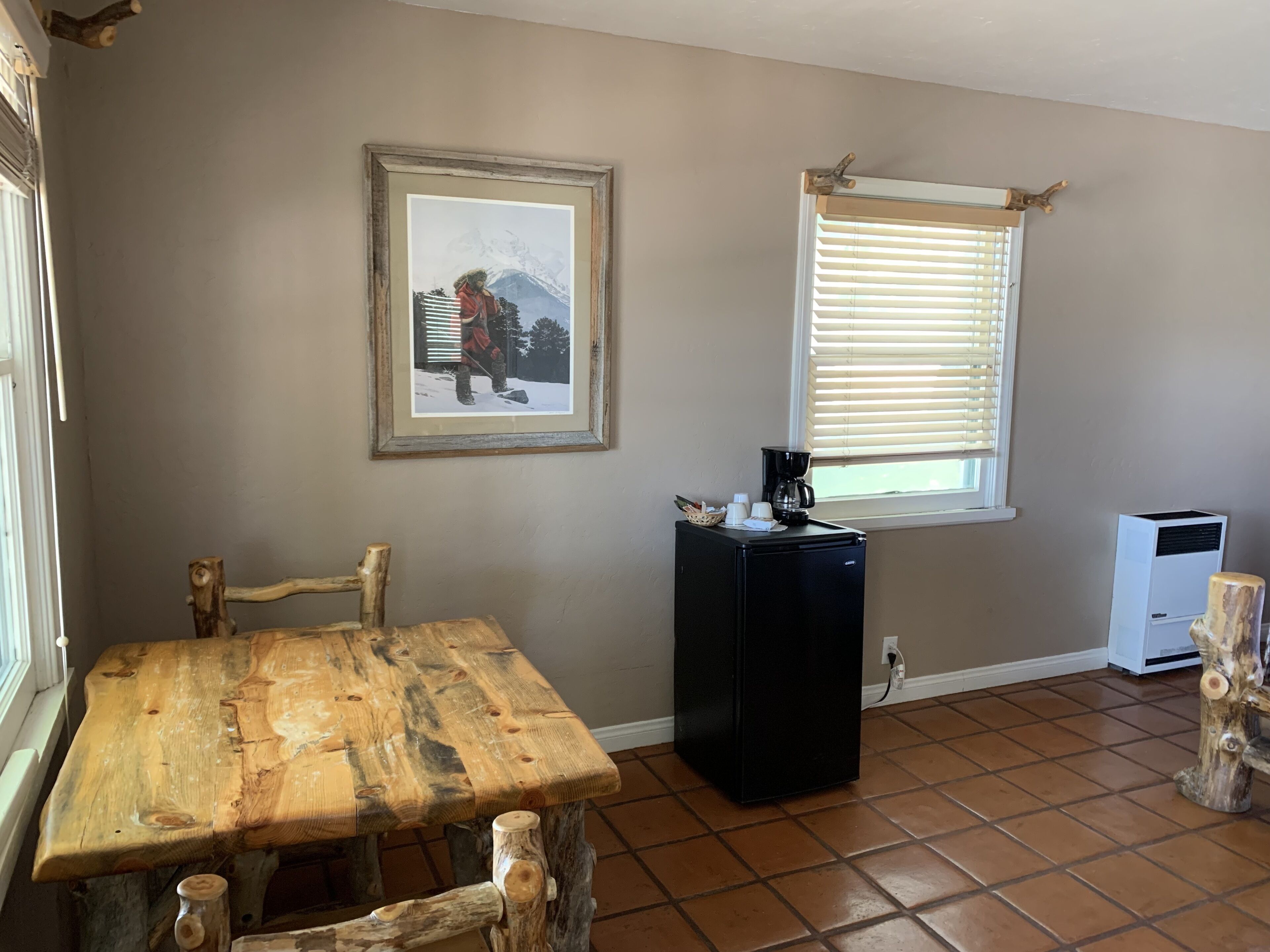 A room with a rustic wooden table, a framed picture on the wall, a window with blinds, a coffee maker, and a small white appliance.
