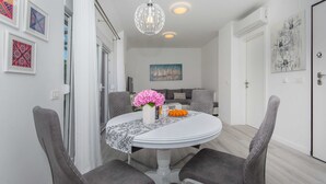 A dining area with a round table, grey upholstered chairs, a white tablecloth, a vase with pink flowers, and a bowl of oranges. - Apartment Gloria (Dubrovnik)