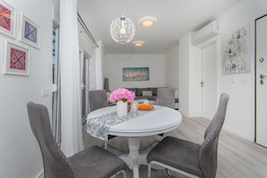 A dining area with a round table, grey upholstered chairs, a white tablecloth, a vase with pink flowers, and a bowl of oranges. - Apartment Gloria (Dubrovnik)