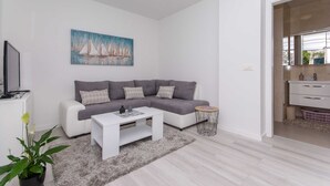 A modern living room with a grey sectional sofa, a white coffee table, a painting of sailboats, a small side table, and a potted plant. - Apartment Gloria (Dubrovnik)