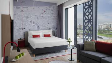 A modern hotel room with a large bed, a sofa, a map on the wall, and a view of the city.